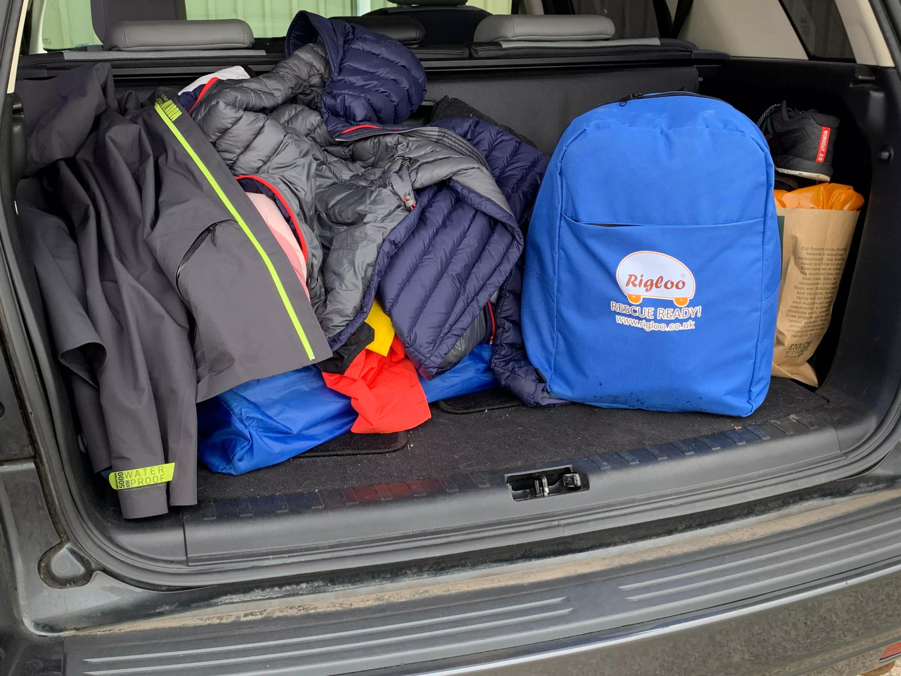 Im Kofferraum eines Autos liegt ein RIGLOO Refuge Notfallzelt samt blauem Rucksack, Kleidung und Wanderschuhen für den sofortigen Einsatz bei Notfällen.