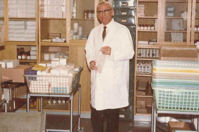 Ein Mann im weißen Laborkittel hält ein kleines Objekt, neben ihm ein Metallwagen mit ordentlich gestapelten Boxen und Behältern.