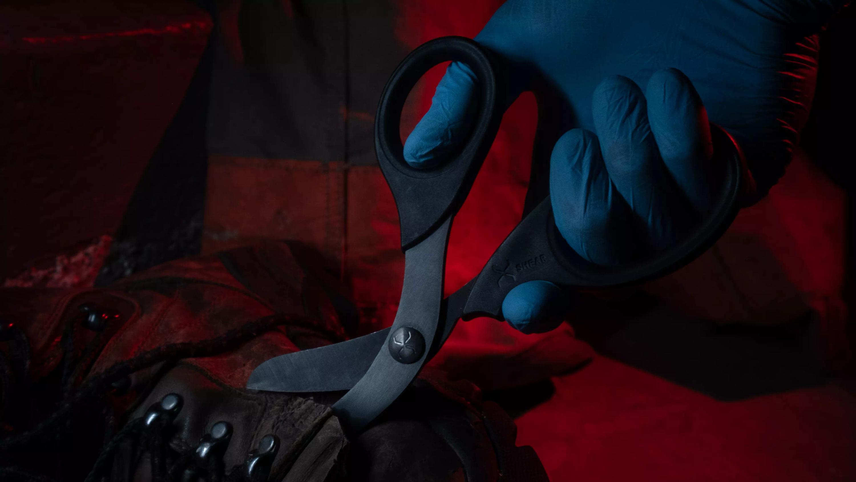 Eine Hand mit blauem Handschuh hält eine Traumaschere XSHEAR® - Schwarz mit gehärteten, schwarzen Titan-Klingen und patentierter stumpfer Spitze über einem Schuh.