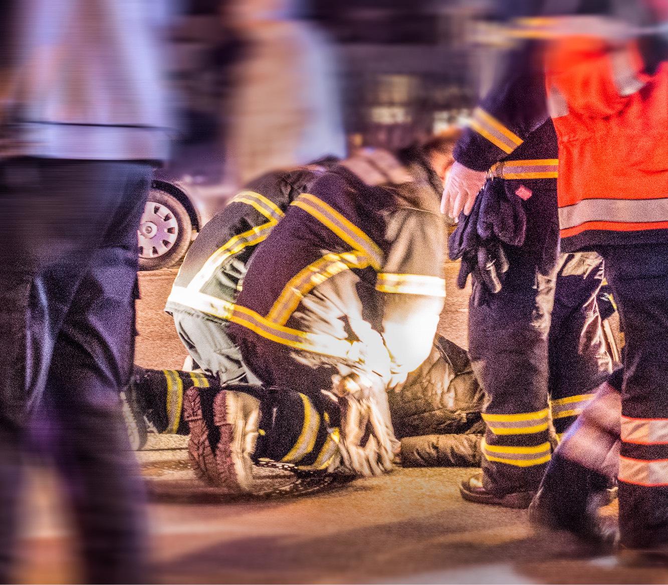 Darstellung eines Unfallszenarios. Eine verunfallte Person liegt auf dem Boden und wird durch zahlreiche Einsatzkräfte versorgt. Im Hintergrund ist ein Auto zu erkennen, das auf dem Dach liegt.