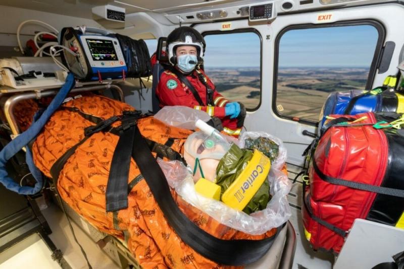 Sanitäter im roten Schutzanzug versorgt bewusstlosen Patienten mit orangefarbener Decke und Beatmungsbeutel auf Trage im Rettungswagen.