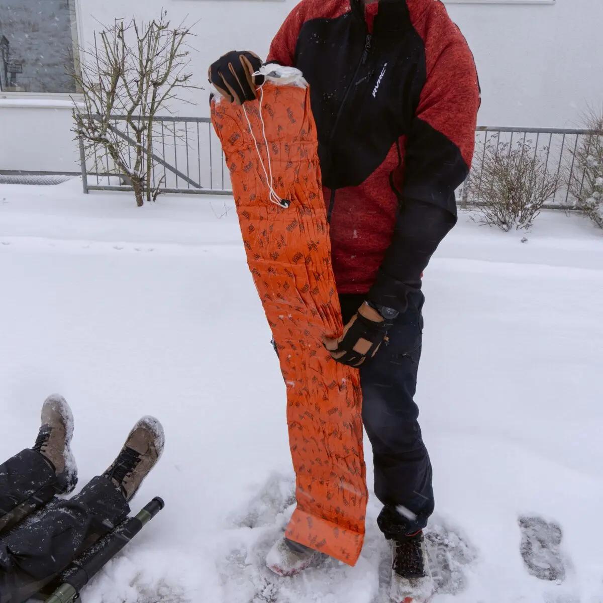 Eine Person hält den Blizzard - Orange, Schlafsack mit Blizzard Reflexcell™ Technologie und bereitet ihn im Schnee zum Einrollen vor.