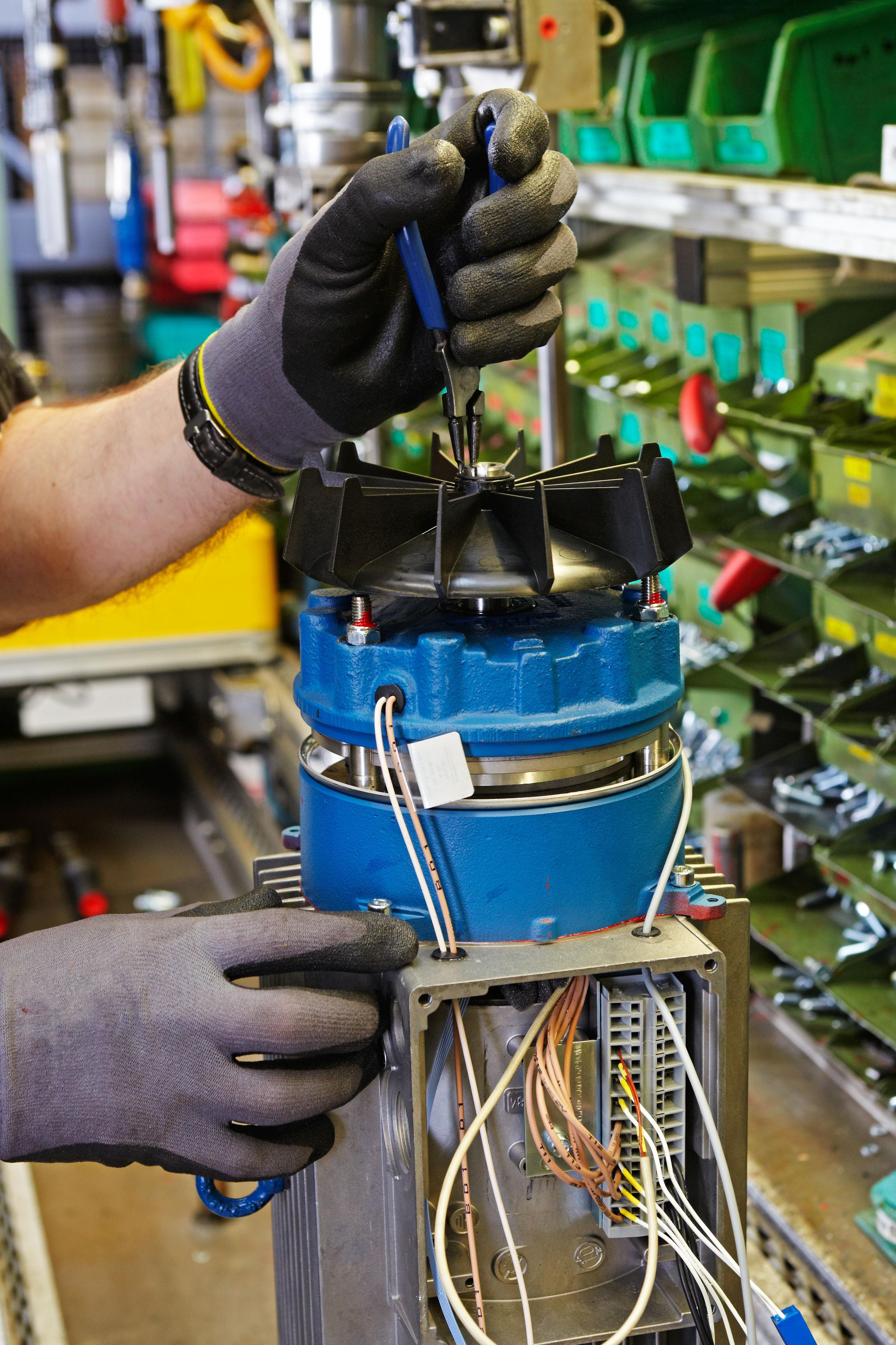 Hand mit schwarzen Arbeitshandschuhen repariert mit blauen Zangen einen verkabelten, blauen Getriebemotor in einer gut ausgestatteten Werkstatt.