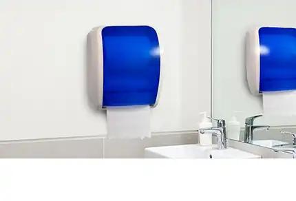 Blauer Papierhandtuchspender aus Kunststoff an weißer Badezimmerwand neben Waschbecken mit silbernem Wasserhahn und weißen Badaccessoires.