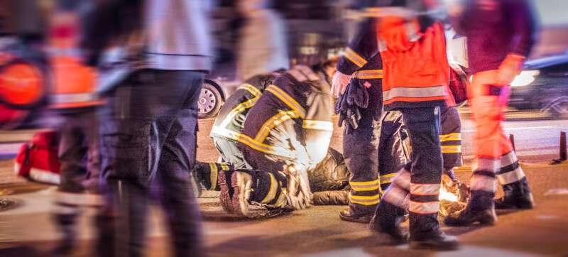 Mehrere Feuerwehrleute mit reflektierenden Streifen leisten nachts Erste Hilfe an einer am Boden liegenden Person neben einem beschädigten Fahrzeug.