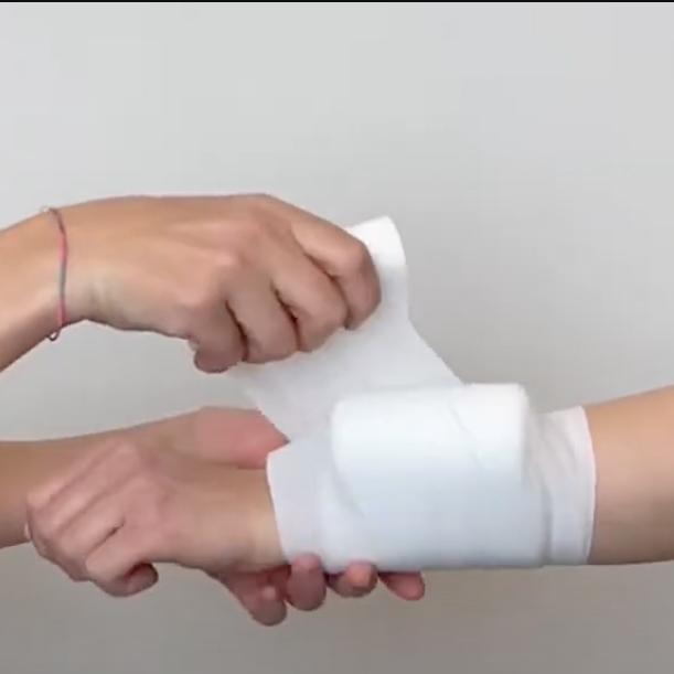 Eine Hand wickelt eine weiße Mullbinde um ein Handgelenk, das von einer farbigen, dünnen Armbandschnur geziert wird.