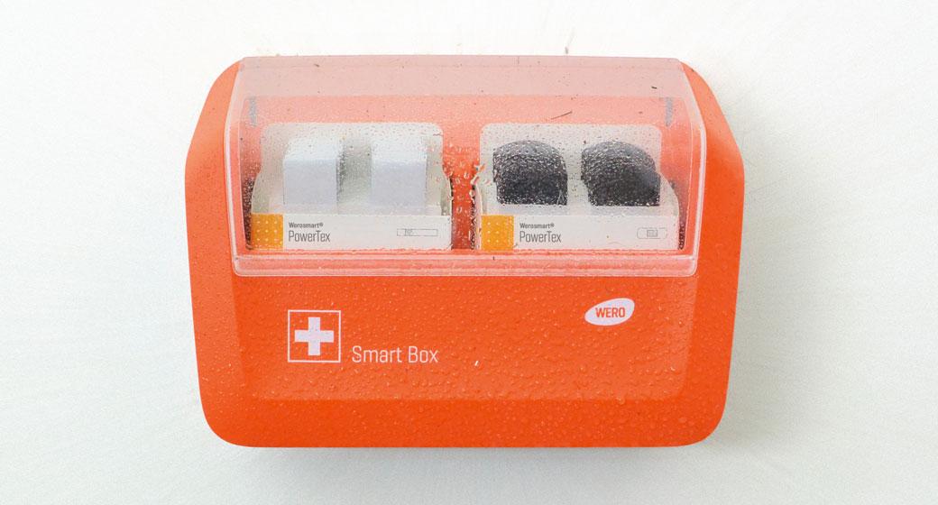 Orangefarbene Wero Smart Box mit PowerTex-Branding, enthält zwei weiße Powerbanks hinter transparenter Abdeckung, mit sichtbaren Wassertropfen.