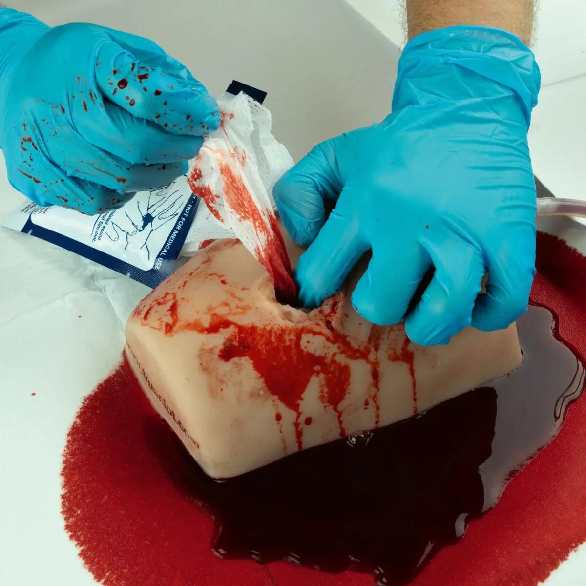 Ein TrueClot® Wound Packing Task Trainer Schusswunden liegt in roter Simulationflüssigkeit, wird mit Schläuchen und Messgerät von zwei blau behandschuhten Händen trainiert.