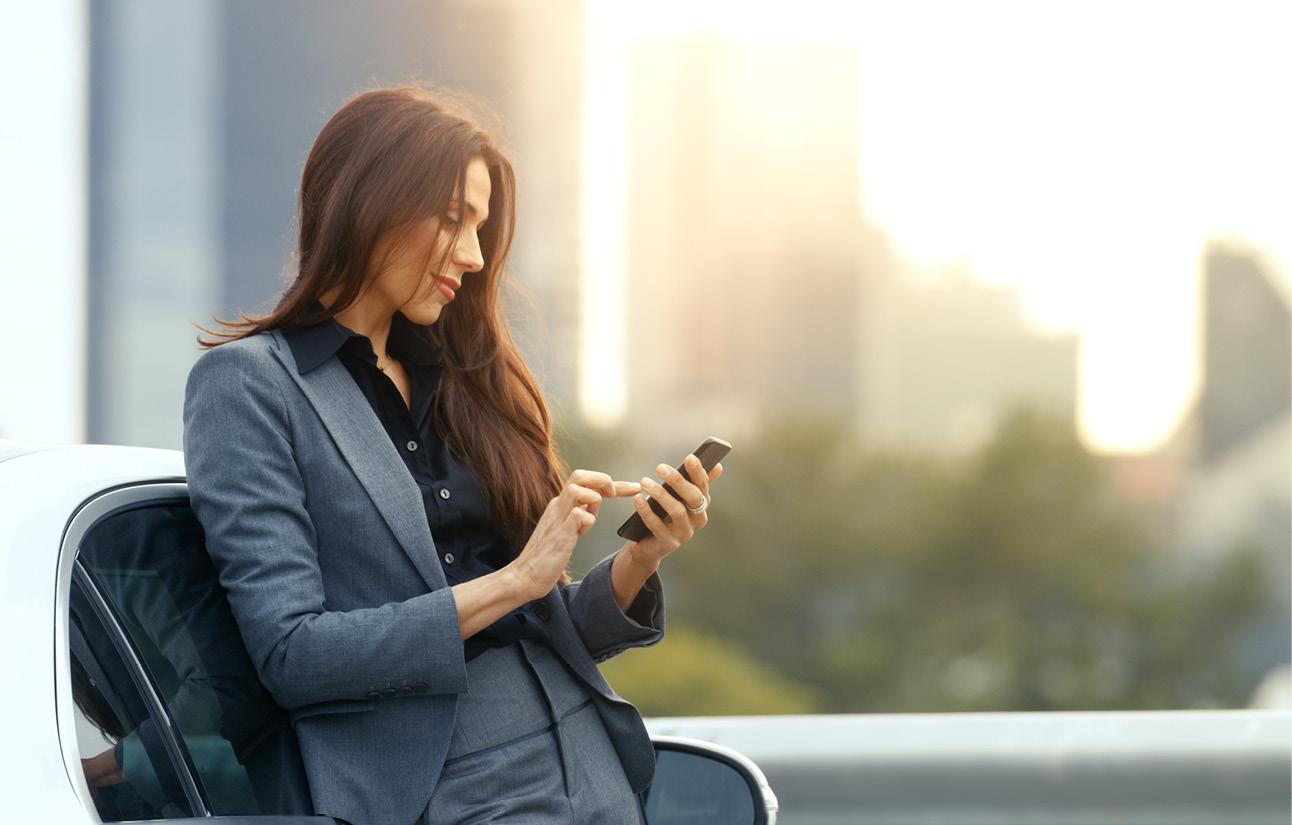 Eine Frau im Business-Outfit lehnt an einem Auto und tippt auf Ihrem Smartphone.