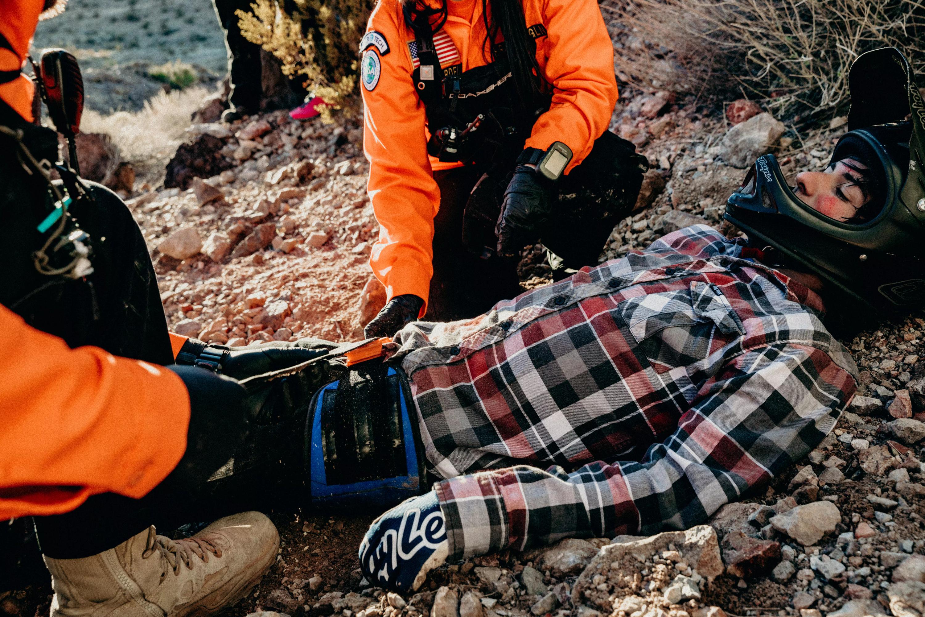 Eine Person erhält in einer Wüstenlandschaft Erste Hilfe, während eine weitere Person den SAM® Pelvic Sling II - Blau, S zur schnellen Versorgung einer Beckenfraktur anlegt.