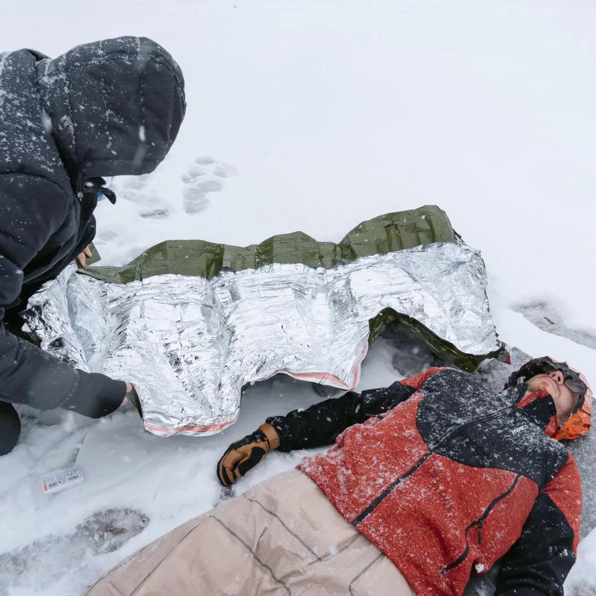 Eine Person in oranger Jacke liegt bewusstlos im Schnee, umhüllt von einer Blizzard - Grün, Decke zur optimalen Wärmespeicherung mit Reflexcell™ Technologie.