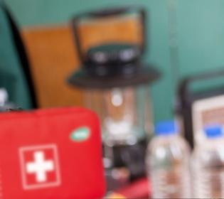 Rotes Erste-Hilfe-Set mit weißem Kreuz, davor eine klare Wasserflasche mit schwarzem Deckel, daneben zwei weitere Flaschen mit blauen Deckeln.