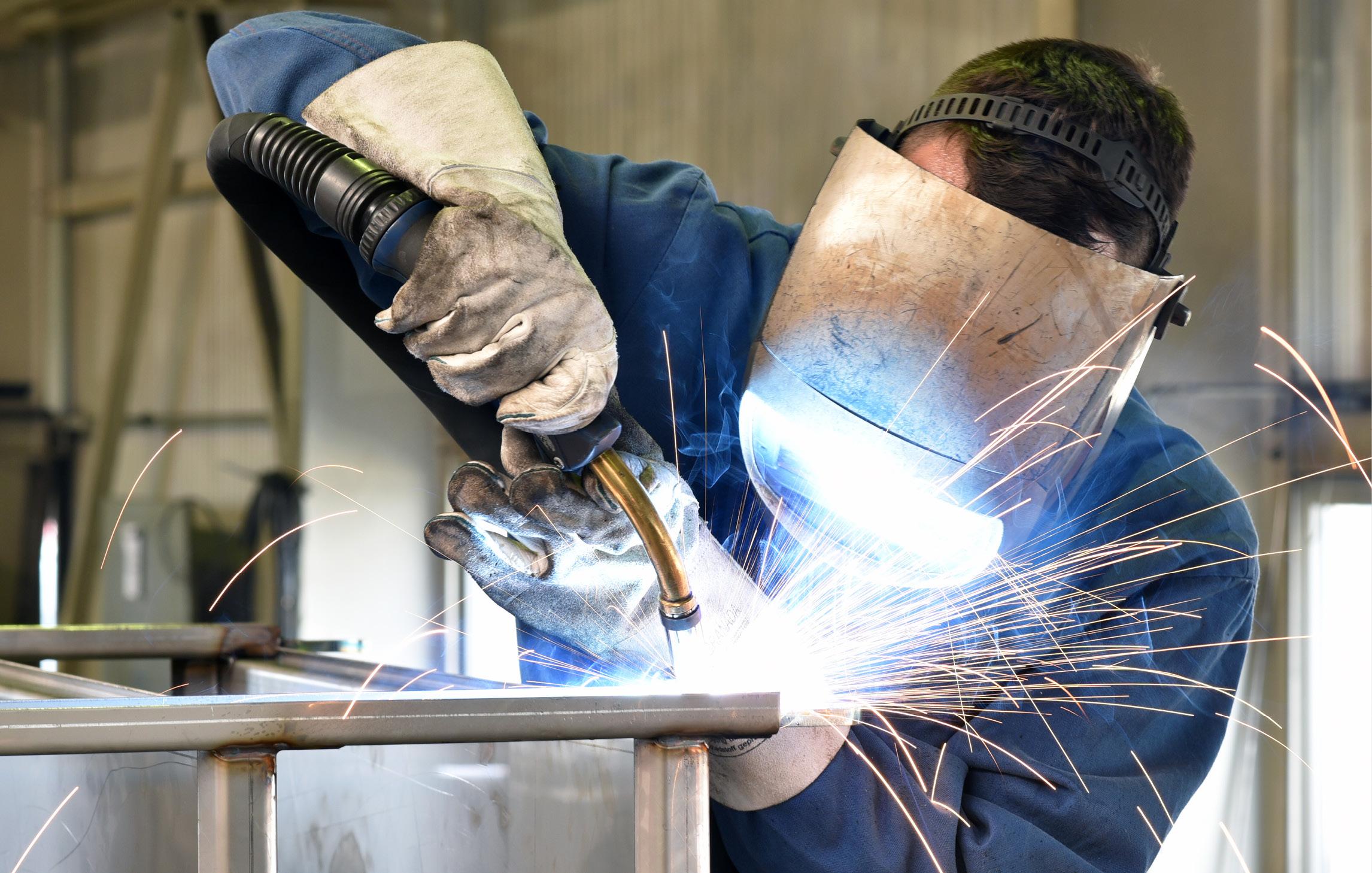 Mitarbeitender schweißt an einem Stück Metall. Funkenflug. Er trägt eine Schweißerbrille und Arbeitshandschuhe
