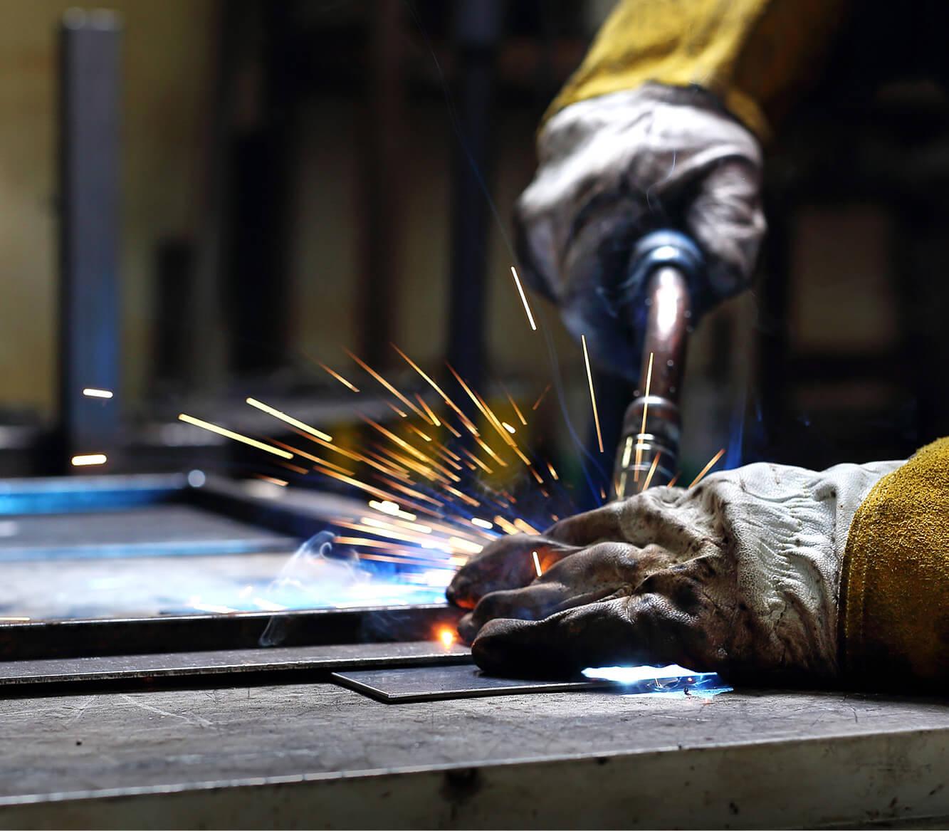 Eine gelbhandschuhte Hand schweißt mit brennendem Schweißbrenner funkenziehend an rauer, dunkler Metalloberfläche.