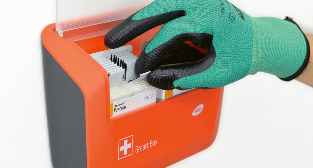 Eine behandschuhte Hand mit schwarzem Fingerschutz greift in einen orangefarbenen, geöffneten Smart Box Erste-Hilfe-Kasten mit PowerTex-Produkten.