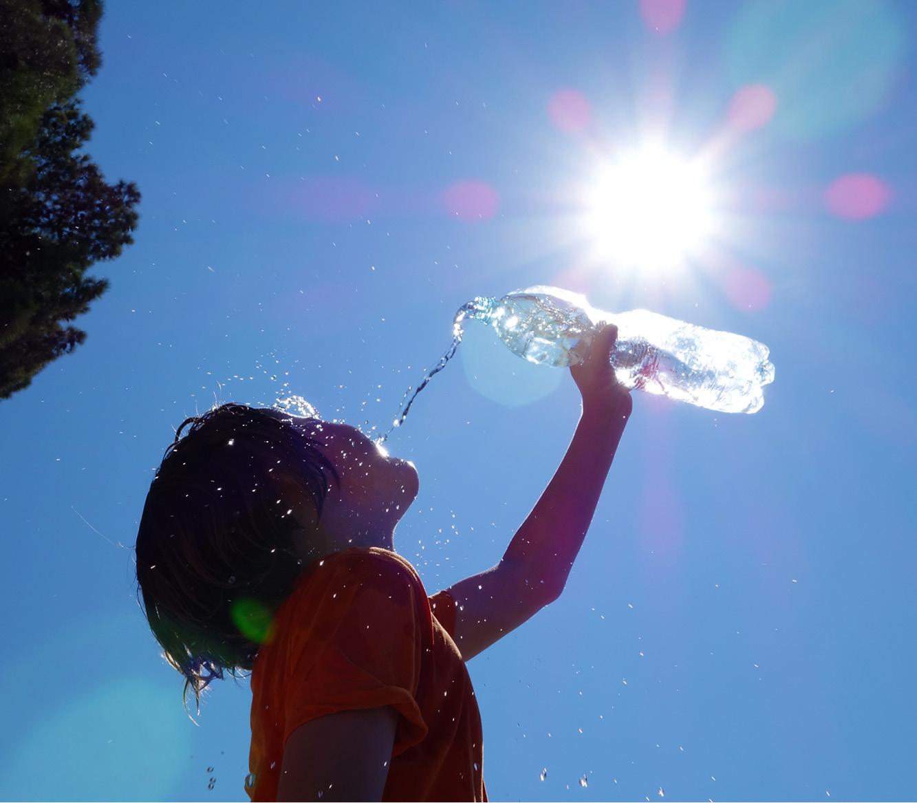 Ein Kind im orangefarbenen T-Shirt trinkt aus einer transparenten Wasserflasche, während Wassertröpfchen vor blauem Himmel spritzen.