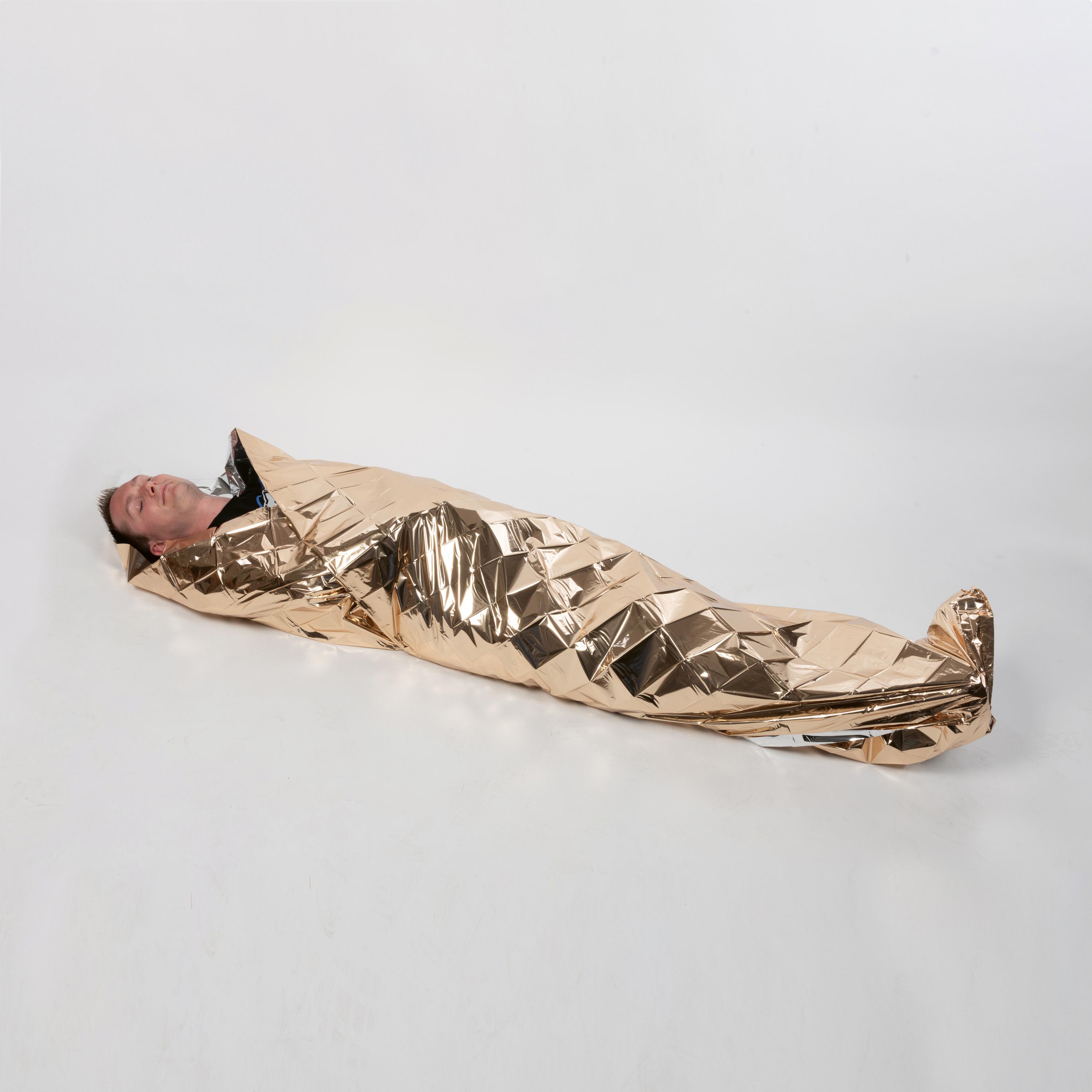 Eine Person liegt in eine goldene WERO Rettungsdecke mit geometrischem Muster gehüllt, die den Körper vollständig bedeckt und Licht reflektiert.