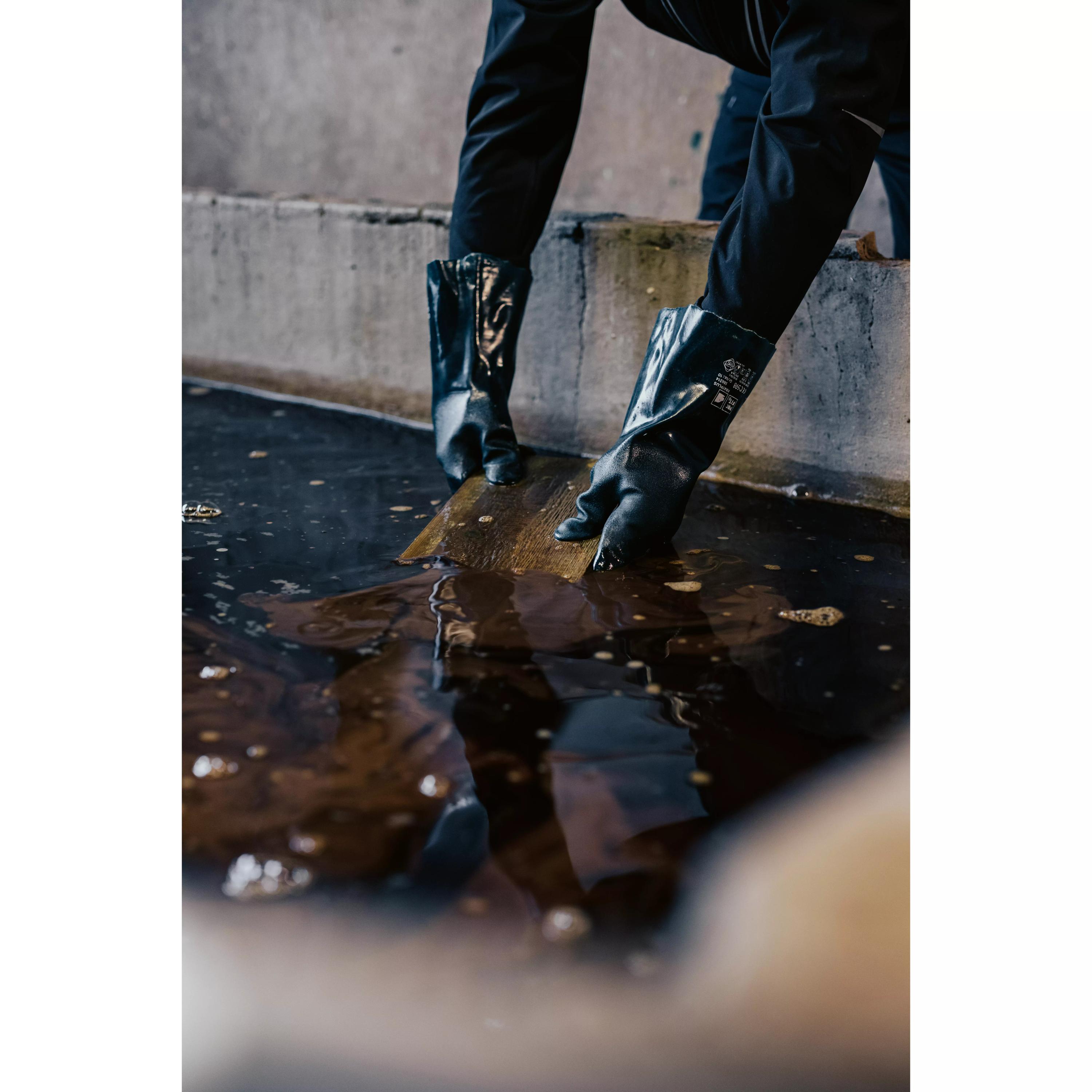 Zwei schwarze Chemikalienschutzhandschuhe Vinyl 40 greifen ein Holzbrett und vermengen damit trübes Wasser, kleine Spritzer betonen die Aktivität.