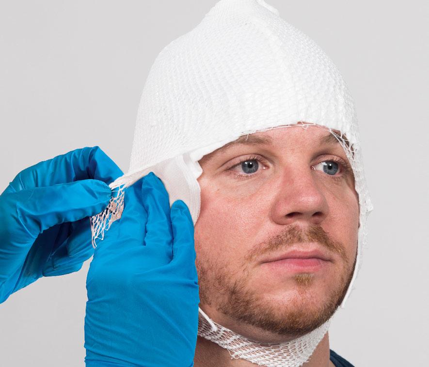 Ein Mann erhält von einer Fachkraft mit blauen Handschuhen eine weiße, netzartige Kopfbandage, die sorgsam angelegt wird.