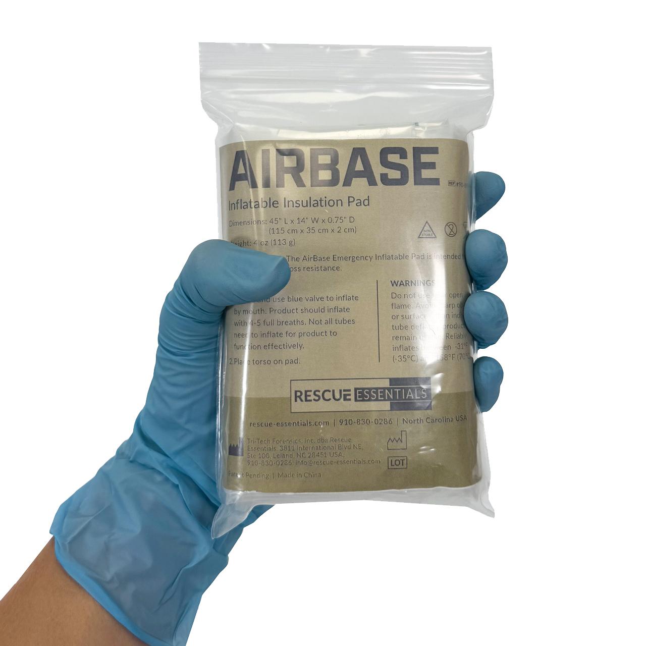 Eine Hand mit blauem Handschuh hält eine Rescue Essentials® AirBase Aufblasbare Isoliermatte im transparenten Beutel mit Produktlabel, kompaktem Design und unabhängigen Luftkammern.