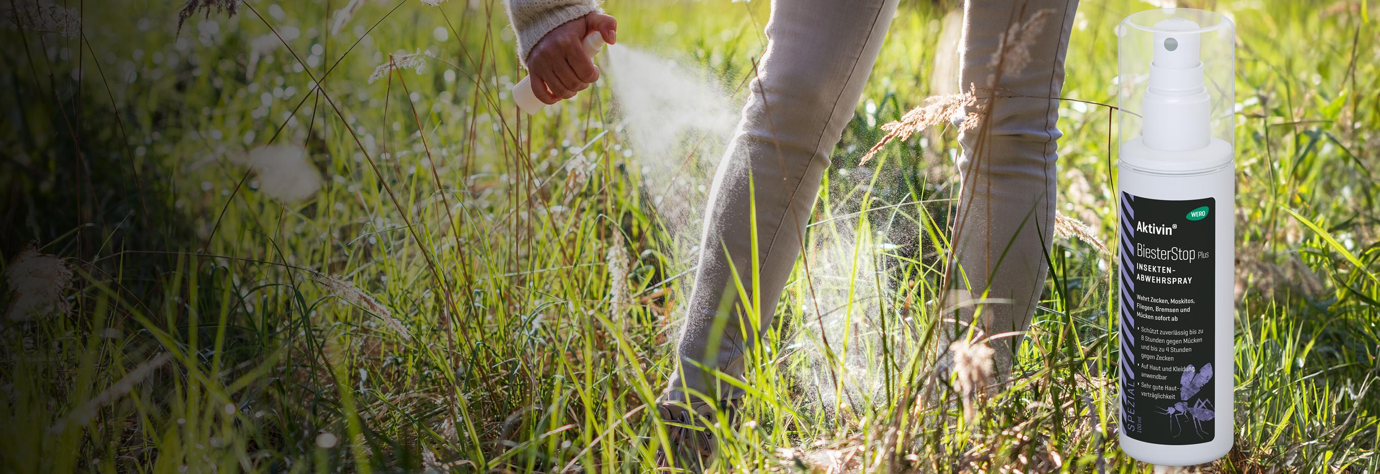 Eine Hand sprüht Akvitin Biester Plus Insekt Abwehrspray aus weißer Flasche mit gelber Flüssigkeit auf hohes, grünes Gras.