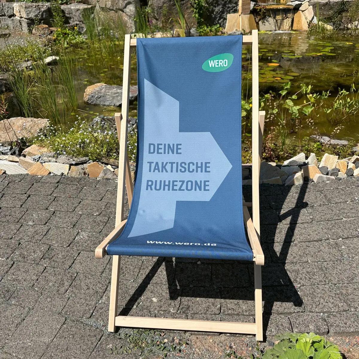 WERO MED-X® Liegestuhl aus robustem Holz mit blauem Bezug, weißem Slogan und WERO-Logo steht auf einem Gartenweg vor Teich und Wasserfall.