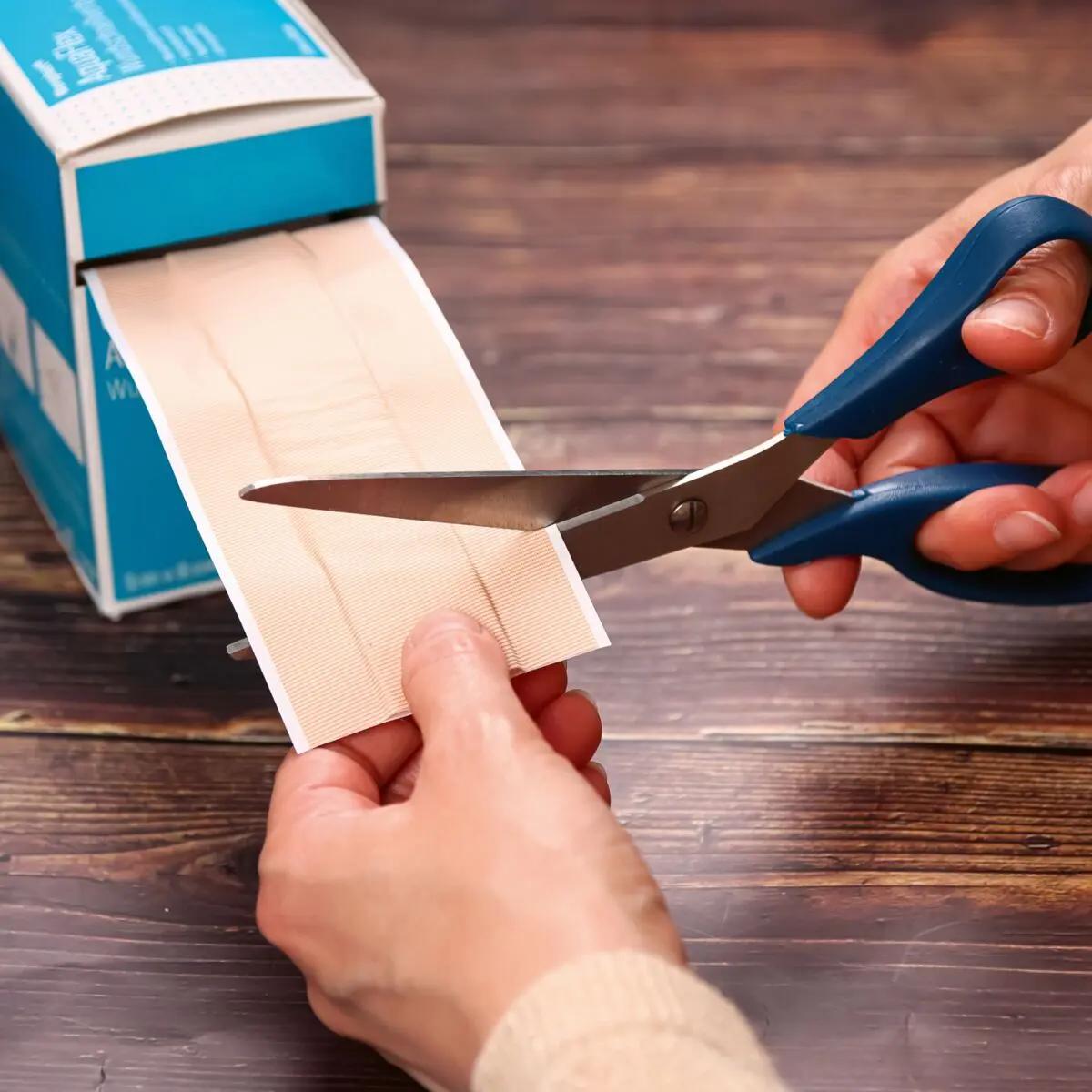 Zwei Hände schneiden mit blauen Scheren einen Streifen des Wundschnellverband Weroplast® AquaFlex, der wasserdicht, superelastisch und flexibel ist.