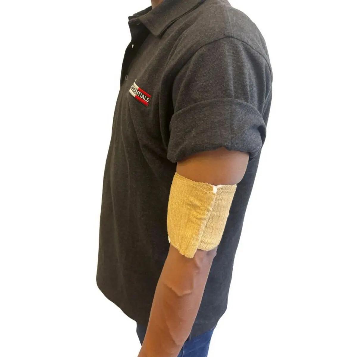 Eine Person trägt die Rescue Essentials® Mini Compression Bandage als beigen Verband mit sichtbarer Falte am Unterarm über einem dunkelgrauen Polo und blauen Jeans.