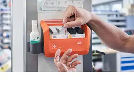 Eine Hand hält ein weißes Tuch über einen geöffneten Erste-Hilfe-Kasten mit Pflasterspender, Gel und Sprayflasche an einer grauen Wand.