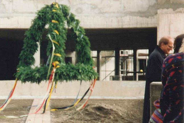 Kranz aus grünen Blättern mit bunten Bändern hängt vor Gebäude im Rohbau mit sichtbarem Gerüst, zwei Personen im Vordergrund.