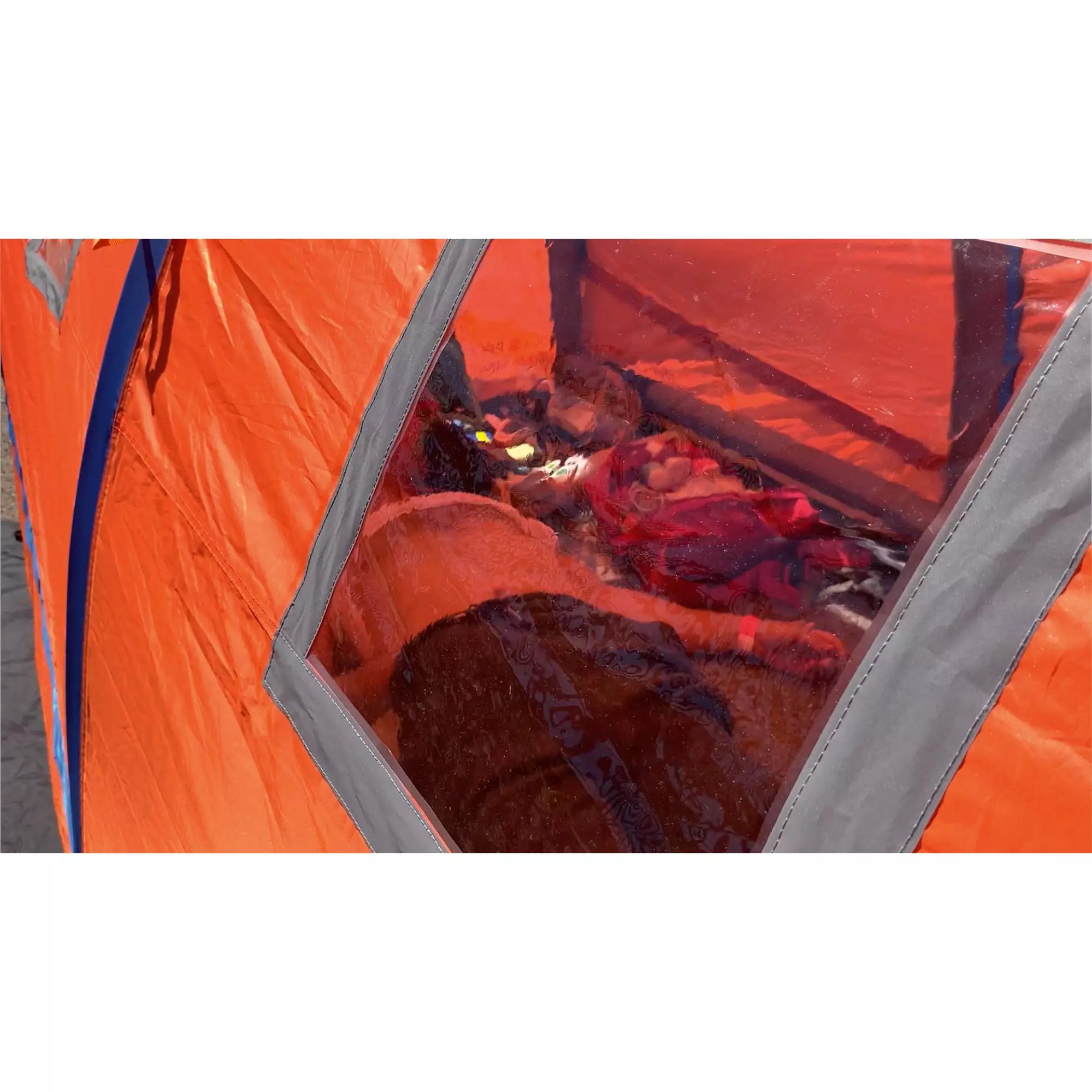 Ein orangefarbenes RIGLOO Rescuer Rettungszelt mit klarem Fenster, stabilem dunklem Rahmen und sichtbarer Person im Inneren auf kiesigem Boden.