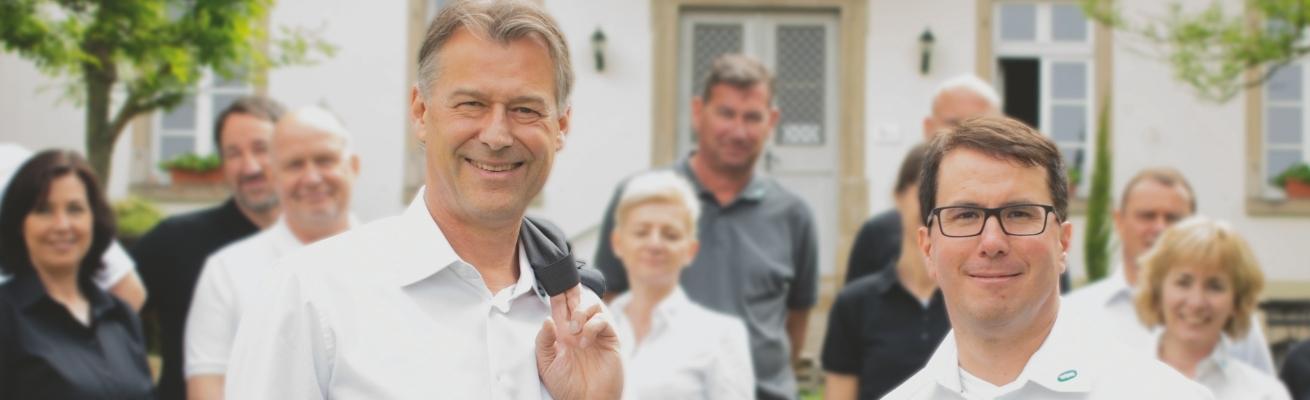 Ein Mann im weißen Hemd mit schwarzer Krawatte steht mit Kollegen in Business Casual Kleidung vor einem weißen Haus im grünen Garten.