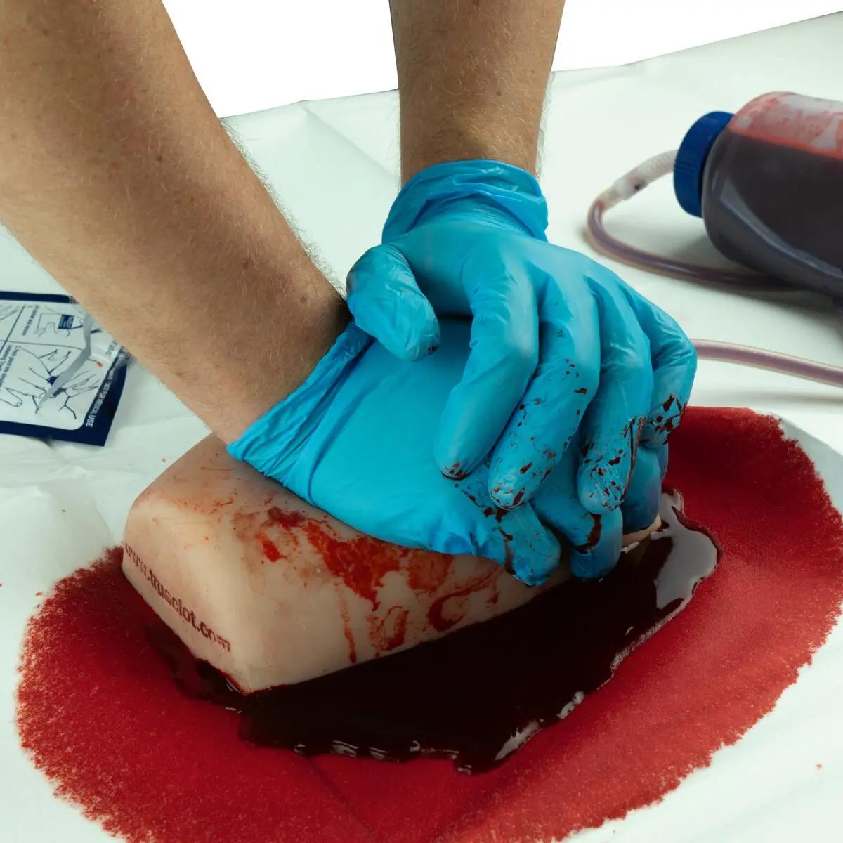 Zwei behandschuhte Hände üben an einem blutverschmierten TrueClot® Woundpacking Trainingssystem - Schusswunde mit weißer Aufhängung eine realistische Blutstillung.