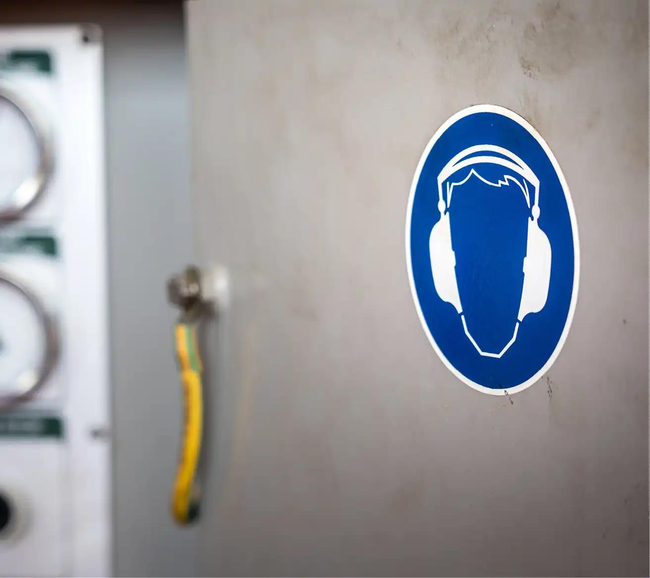 Blaues Sicherheitsschild mit weißem Kopfhörersymbol auf grauer Maschinentür.
