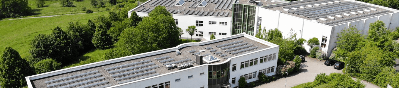 Moderner Gebäudekomplex mit weißer Fassade, geschwungener Glasfront, Solarpanels auf den Dächern und angrenzenden Parkflächen.