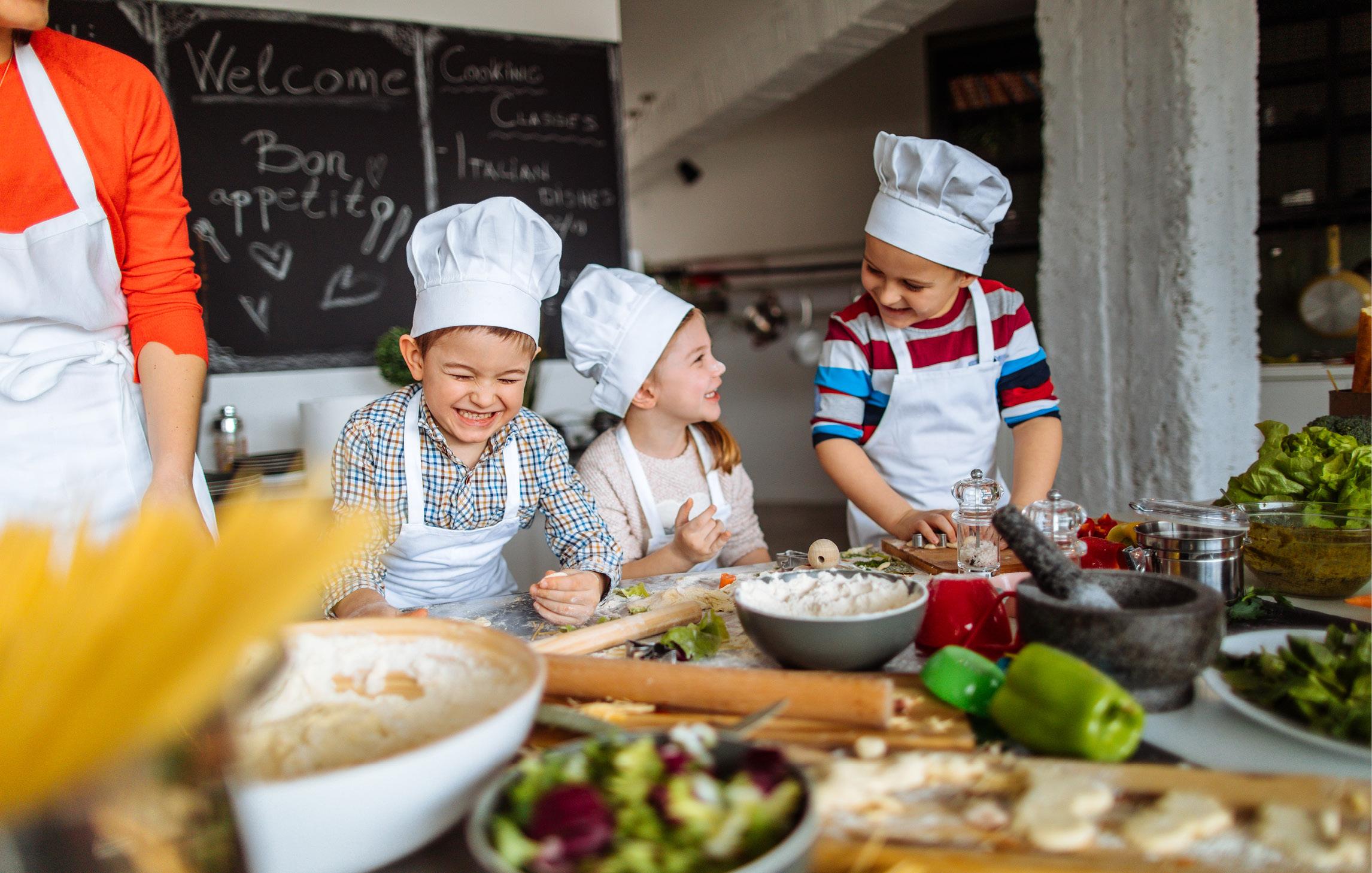 Drei Kinder stehen an einer niedrigen Arbeitsplatte in einer Kindertagesstätte und kochen. Sie tragen Schürzen und kleine Kochmützen