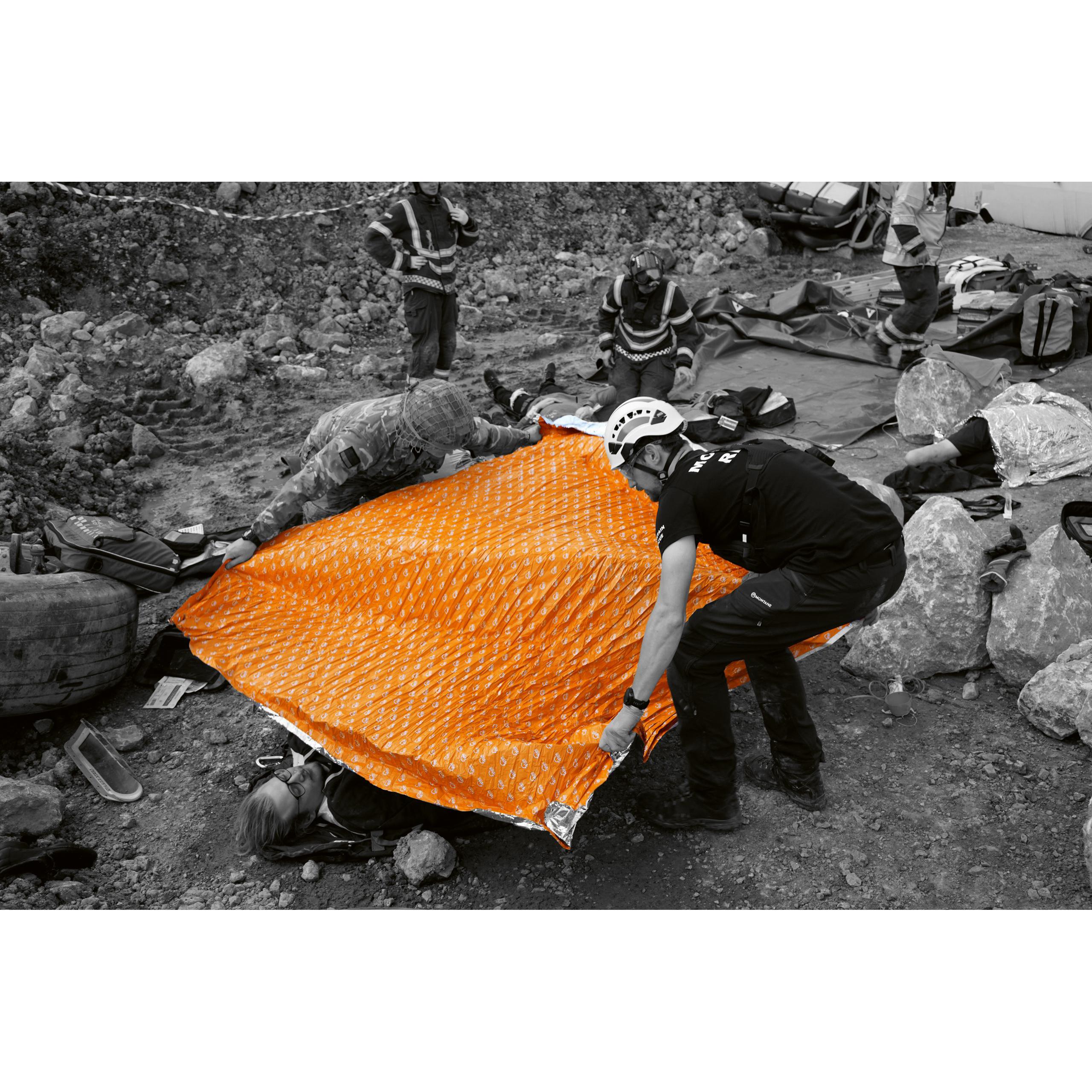 Eine Person legt auf felsigem Untergrund eine orange igloo Survival Pro - Orange, Größe L Rettungsplane aus, weitere Menschen agieren im Hintergrund.