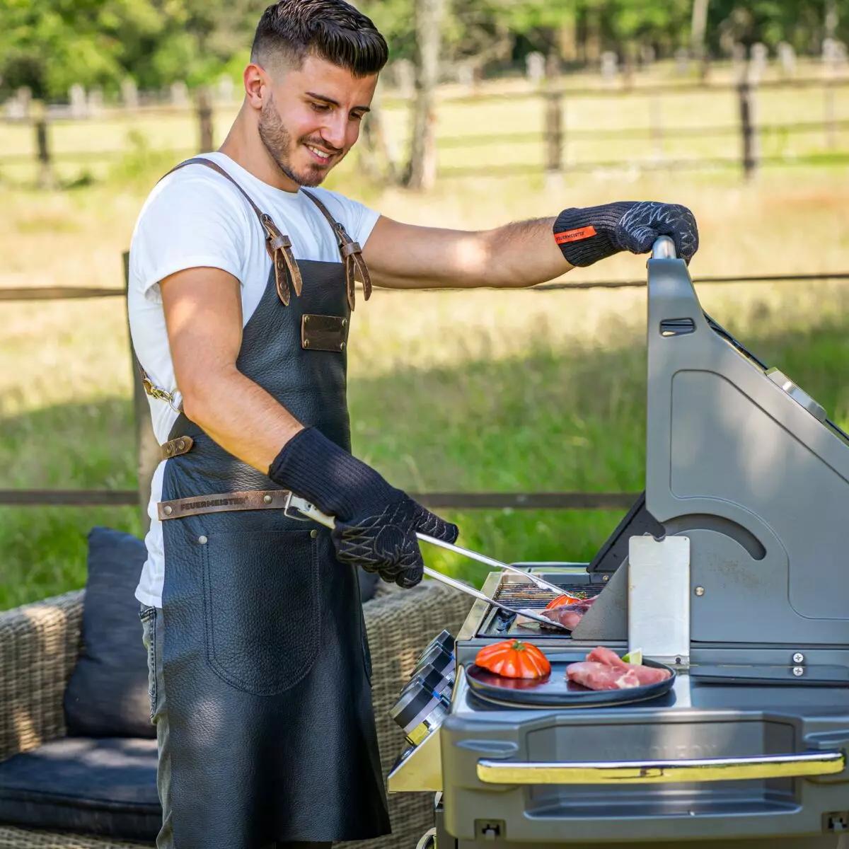 Ein junger Mann grillt Fleisch an einem großen Grill und trägt dabei Hitzeschutzhandschuh Aramid, 5 Paar - 8, geeignet für hohe Temperaturen.