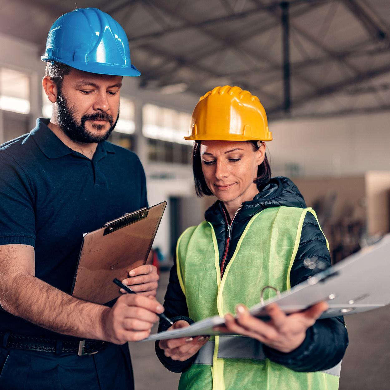 Zwei Menschen mit Schutzhelm stehen nebeneinander. Eine Person hält einen Ornder, während die andere mit einem Stift etwas im Ordner aufschreibt.