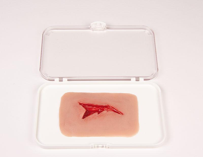 Eine Erler-Zimmer Woundmoulage Risswunde liegt in einer weißen Kunststoffschale, das Modell zeigt einen unregelmäßigen, blutenden Riss in hellrosa Schaumstoff.