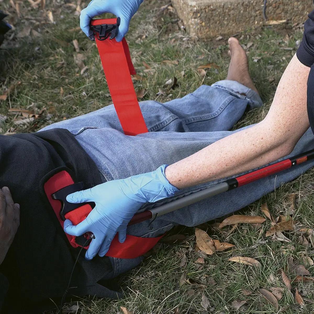 Eine Person mit blauem Handschuh trägt eine Slishman Traction Splint (STS) GEN. 2 am Bein, roter Gurt an der Hüfte und Stützstange stabilisieren das Bein.