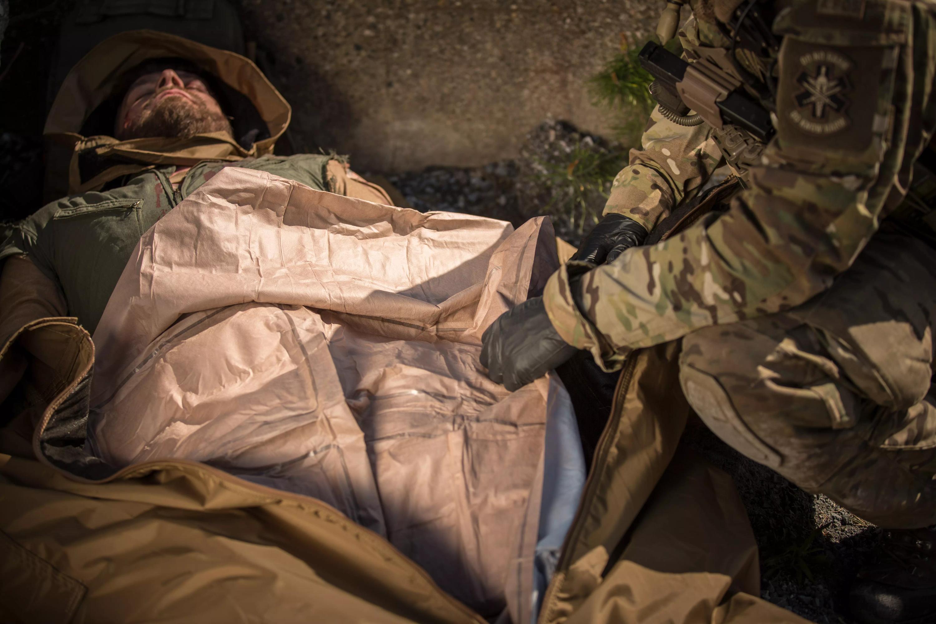 Eine Person liegt im Freien und wird von einer Ready-Heat™ - 6-Element-Decke in Beige umhüllt, während jemand in Militärkleidung helfend daneben kniet.
