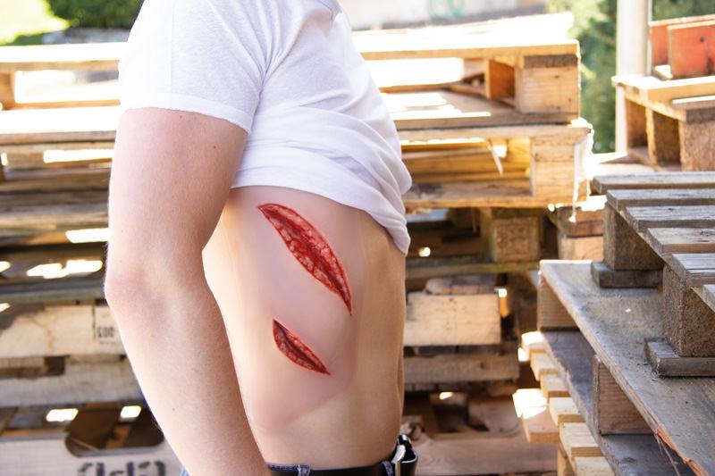 Eine Person trägt eine Erler-Zimmer Woundmoulage Schnittwunde groß mit Blutungsfunktion am seitlichen Oberkörper, die realistisch eine tiefe, blutige Verletzung darstellt.
