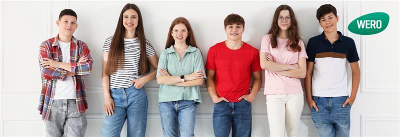 Sechs Teenager mit unterschiedlicher Kleidung stehen nebeneinander vor einer weißen Wand, alle schauen nach vorn, einige mit verschränkten Armen.