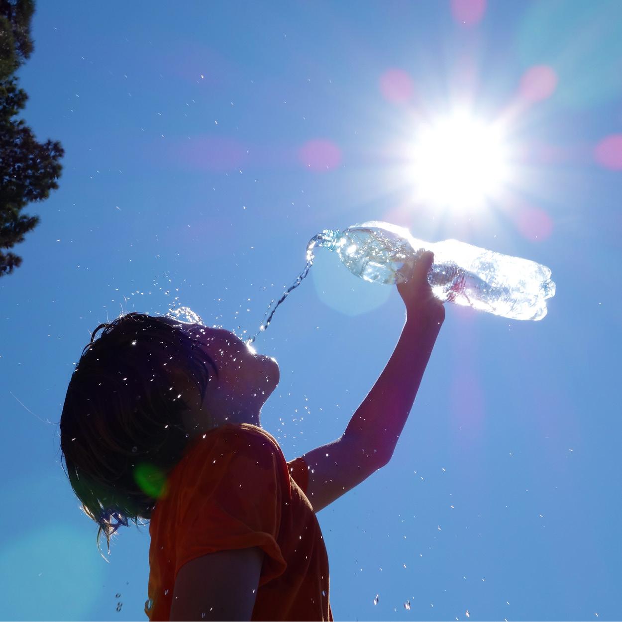 Ein Kind guckt in Richtung Sonne. Zur Abkühlung gießt er eine Flasche Wasser über sich.
