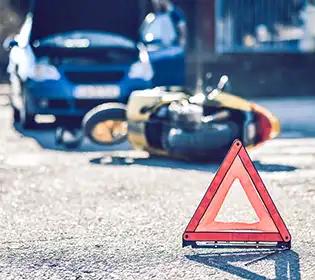 Gelbes Motorrad liegt umgestürzt auf Asphalt neben rotem Warndreieck, im Hintergrund steht ein Auto mit offener Motorhaube.