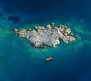 Ein orange-rotes Kajak treibt auf türkisfarbenem Wasser neben einer felsigen, grau-weißen Insel aus der Vogelperspektive.