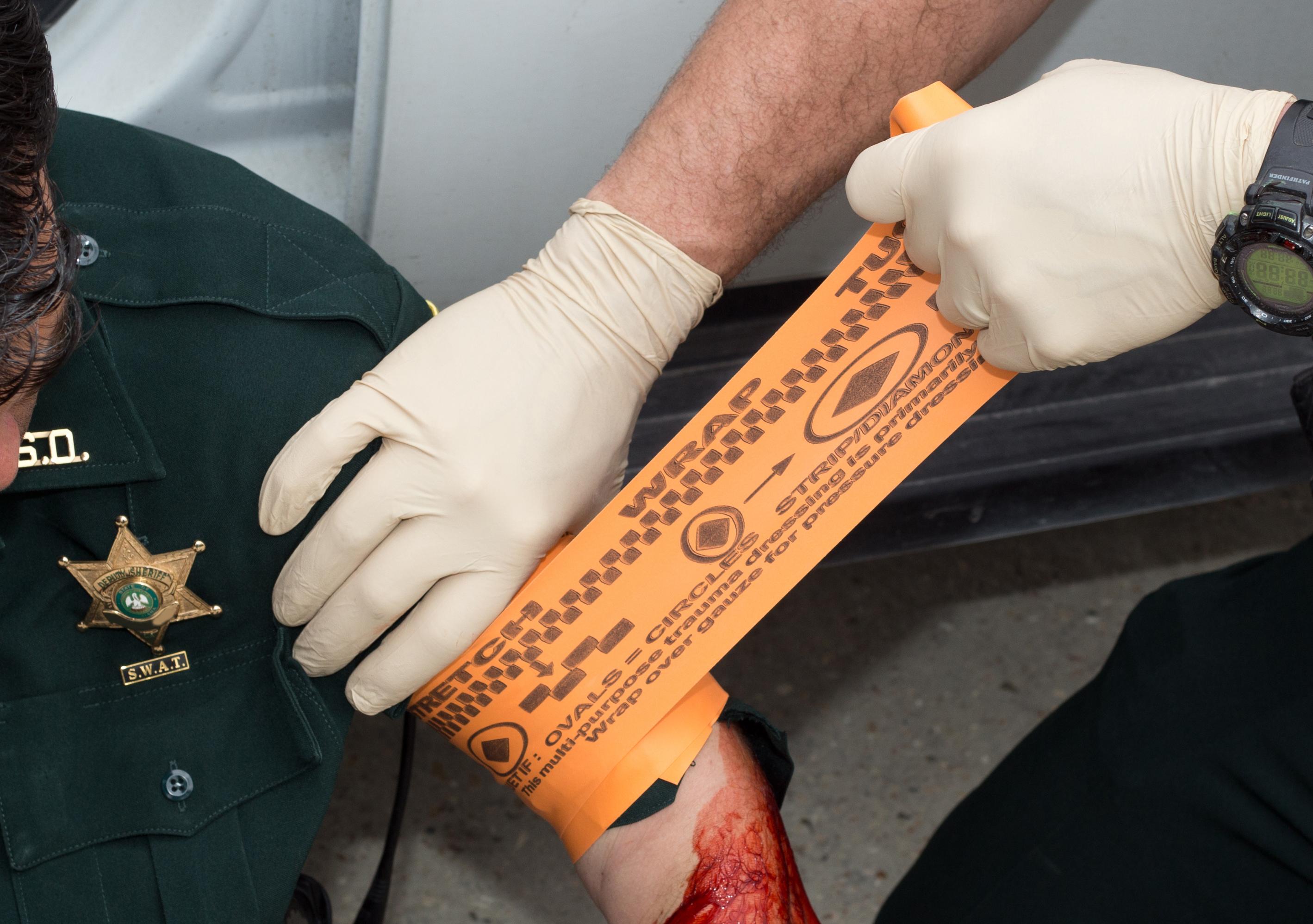 Ein Polizist mit grünem Uniformärmel wickelt ein Tourniquet SWAT - T - Orange mit gut sichtbarer Anleitung um seinen Unterarm.