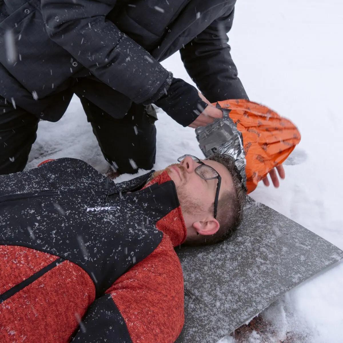 Eine Person erhält auf schneebedecktem Boden Erste Hilfe; eine zweite Person legt ihr eine Blizzard - Orange, Beanie Mütze mit Blizzard Reflexcell™ Technologie an.