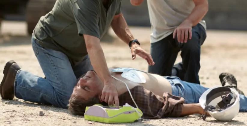 Zwei Personen leisten Erste Hilfe mit einem tragbaren AED und einer CPR-Hilfe bei einer auf dem Boden liegenden Person in Arbeitskleidung.